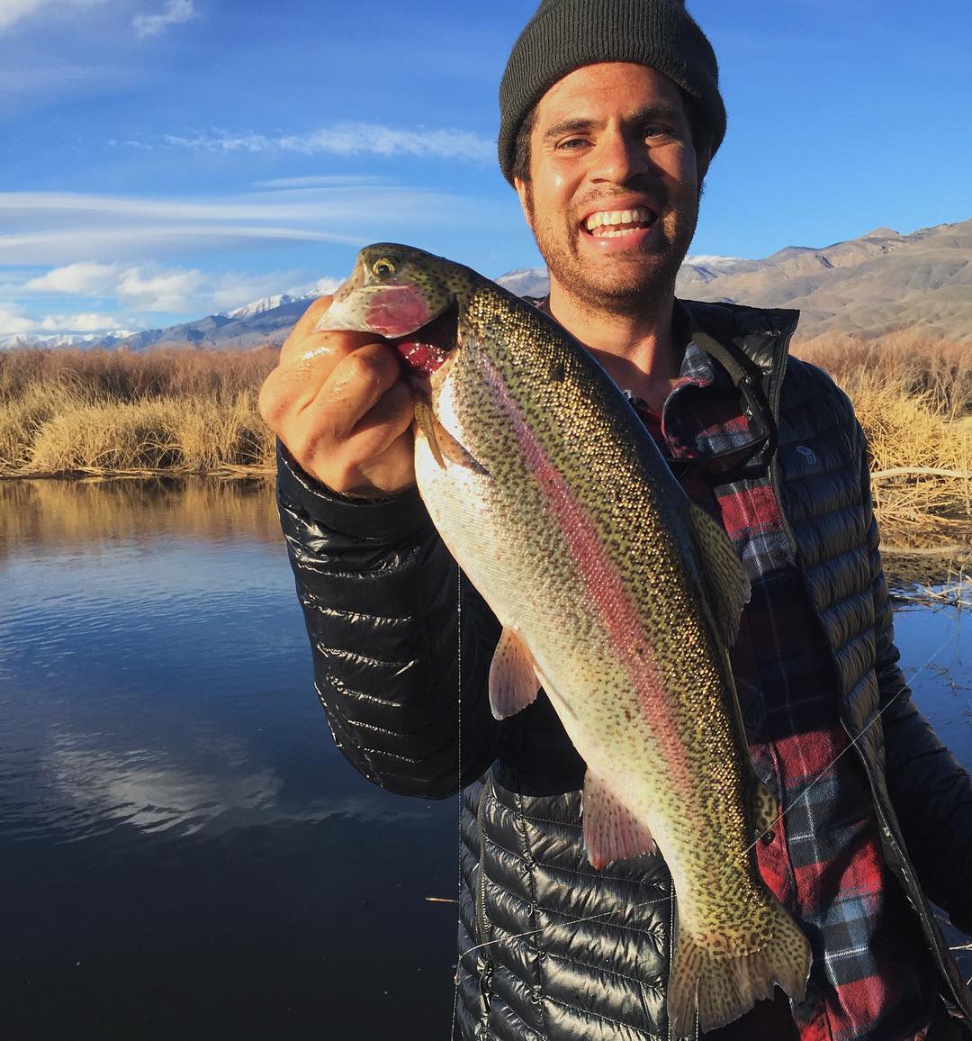 Person smiling and holding a large trout near a river with tall grass and the majestic mountains of Bishop, California in the background. visit bishop