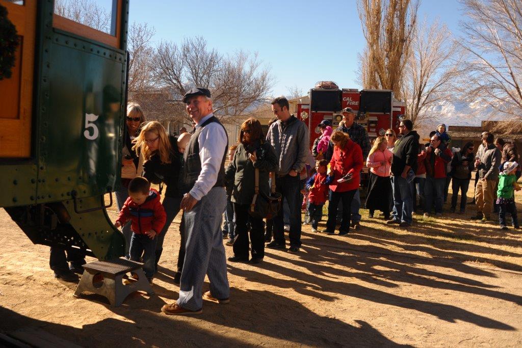 A group of people, including excited kids, line up outside a vintage railroad express on a sunny holiday. visit bishop