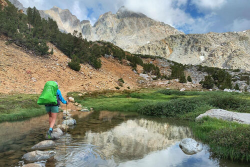 A hiker with a green backpack traverses stepping stones over a serene creek in Bishop, California, with the rugged mountains of the Eastern Sierra looming in the background. visit bishop