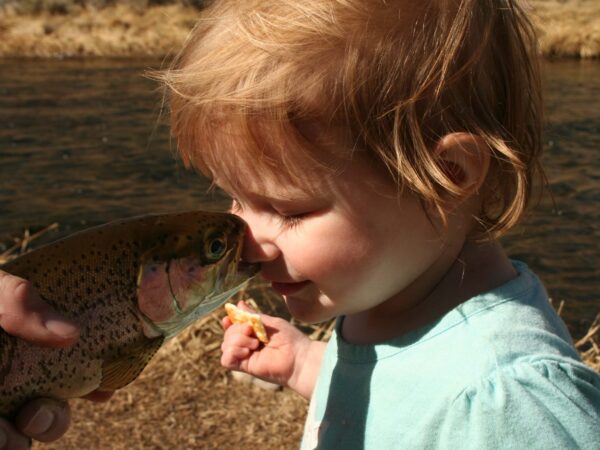 A toddler is gently touching noses with a fish held by an adult near a riverbank. visit bishop