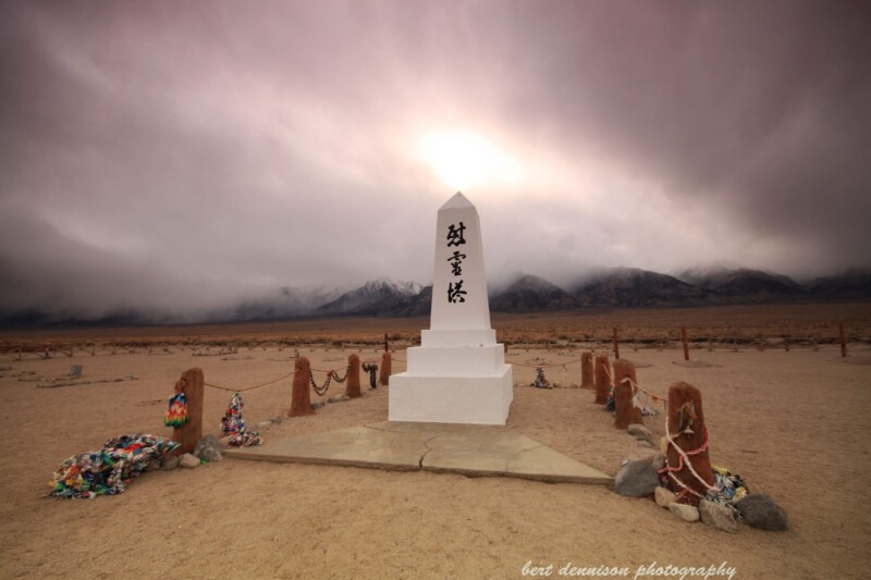 A white obelisk monument stands in a desolate, sandy landscape under a cloudy, dramatic sky in Bishop, California. Colorful fabric tributes and stones are placed around its base. Mountains shrouded in mist appear in the background. The image shows "bert dennison photography" at the bottom. visit bishop