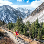 A trail runner, wearing a red outfit, runs along a mountain path with trees in the foreground and snow-capped peaks behind. visit bishop
