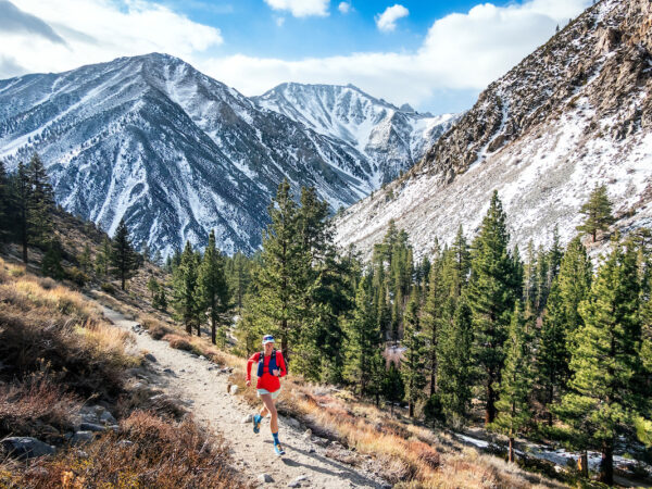 A trail runner, wearing a red outfit, runs along a mountain path with trees in the foreground and snow-capped peaks behind. visit bishop