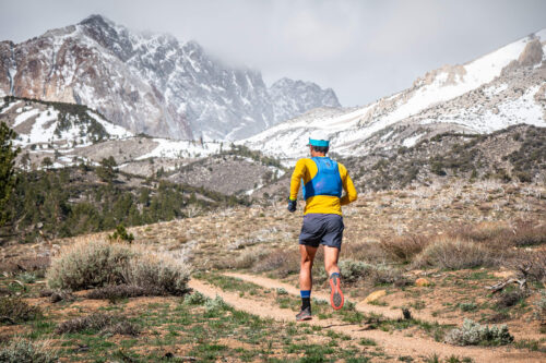 A runner in blue and yellow attire is seen from behind, running on a trail through the rugged mountain landscape of Bishop, California. Snow-capped peaks rise in the background, while the foreground features rocky terrain with patches of grass and shrubs under a cloudy sky. visit bishop