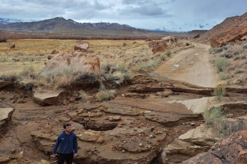 A person stands in a rocky, eroded landscape with mountains in the background under a cloudy sky, capturing the rugged beauty of Bishop, California. visit bishop