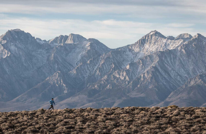 A person walks a dog on a grassy ridge near Bishop, California, with a backdrop of towering, snow-capped mountains under a cloudy sky. visit bishop