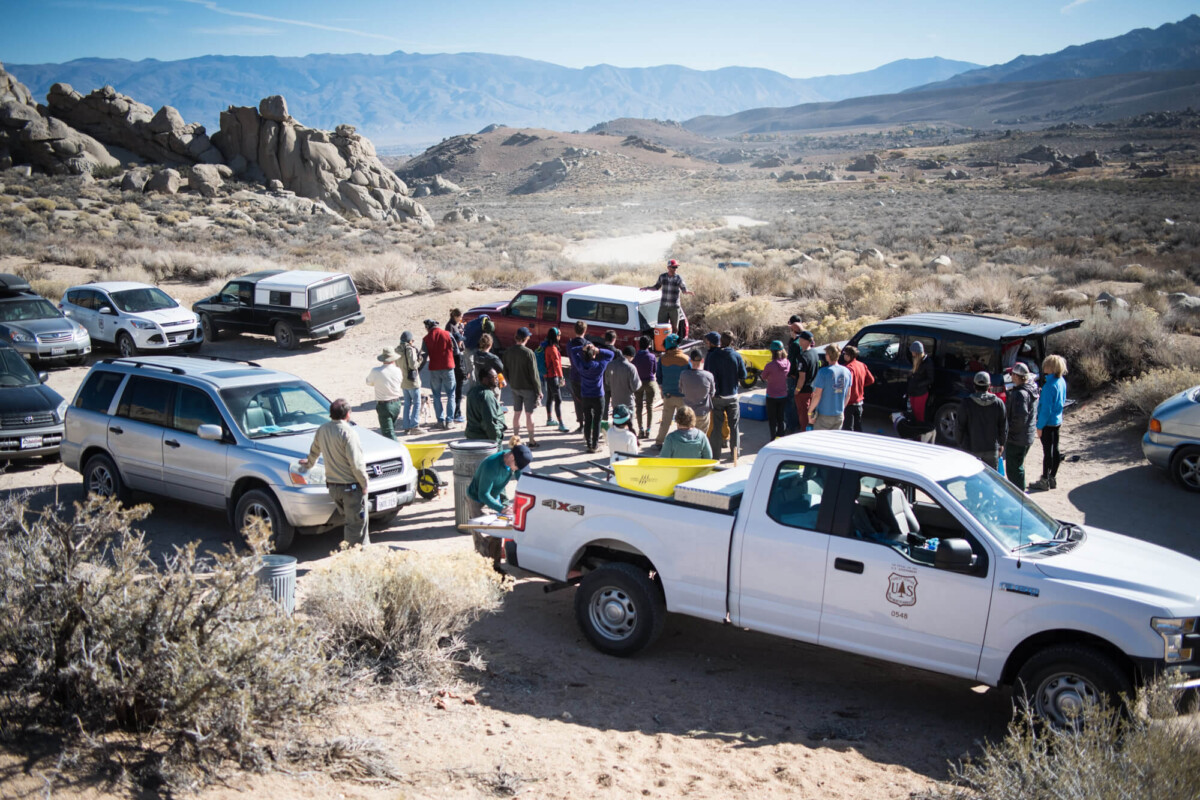 A group of people gathered outdoors in a desert-like area near several parked vehicles, including a white pickup truck with a government emblem. The scene, reminiscent of the rugged landscapes near Bishop, California, features rocky hills and distant mountains under a clear blue sky. visit bishop