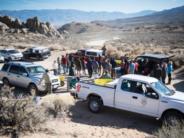 A group of people gathered outdoors in a desert-like area near several parked vehicles, including a white pickup truck with a government emblem. The scene, reminiscent of the rugged landscapes near Bishop, California, features rocky hills and distant mountains under a clear blue sky. visit bishop