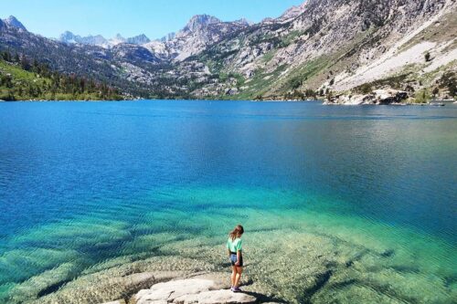 A person stands on a rock at the edge of a clear turquoise lake near Bishop, California, surrounded by mountains with green and rocky slopes under a bright blue sky. The water is so clear that the lakebed is visible near the shore. visit bishop