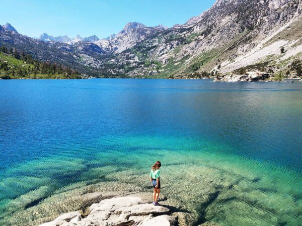 A person stands on a rock at the edge of a clear turquoise lake near Bishop, California, surrounded by mountains with green and rocky slopes under a bright blue sky. The water is so clear that the lakebed is visible near the shore. visit bishop