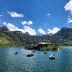 A serene mountain lake with boats anchored, surrounded by the green hills of Bishop, California, and snow-capped peaks of the Eastern Sierra under a blue sky with clouds. visit bishop