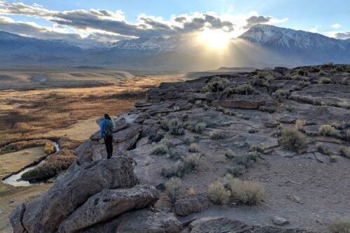 Person standing on a rocky cliff overlooking a vast landscape with mountains and the sun shining through clouds. visit bishop