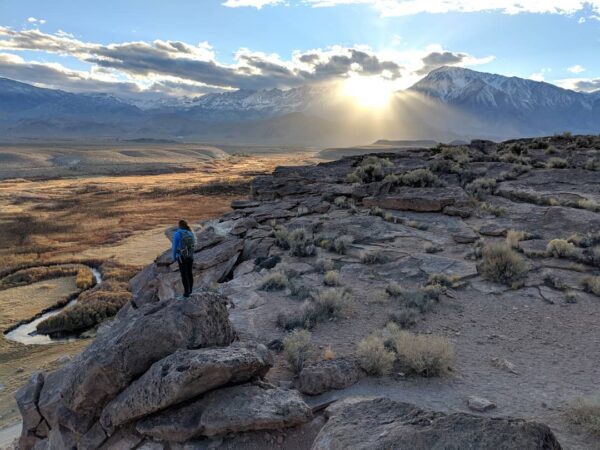 Person standing on a rocky cliff overlooking a vast landscape with mountains and the sun shining through clouds. visit bishop