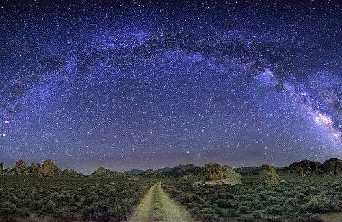 A breathtaking night landscape near Bishop, California, shows the Milky Way galaxy arcing across a clear, star-filled sky. Below the galaxy, a dirt path stretches through a sparse, rocky desert terrain with low vegetation and distant mountains, creating a serene and otherworldly scene. visit bishop