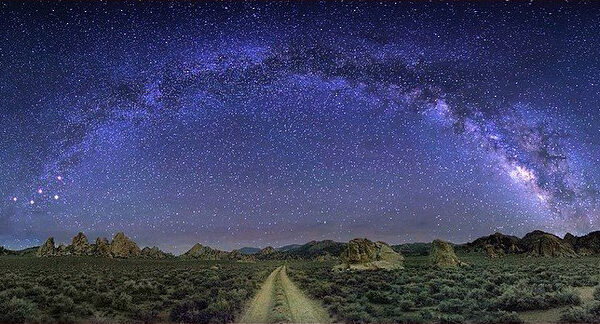 A breathtaking night landscape near Bishop, California, shows the Milky Way galaxy arcing across a clear, star-filled sky. Below the galaxy, a dirt path stretches through a sparse, rocky desert terrain with low vegetation and distant mountains, creating a serene and otherworldly scene. visit bishop