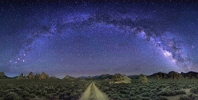 A breathtaking night landscape near Bishop, California, shows the Milky Way galaxy arcing across a clear, star-filled sky. Below the galaxy, a dirt path stretches through a sparse, rocky desert terrain with low vegetation and distant mountains, creating a serene and otherworldly scene. visit bishop