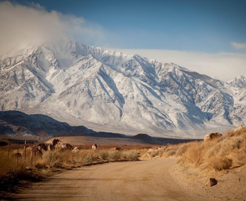 Dirt road leading to snow-capped mountains under a clear blue sky with rocky terrain and dry vegetation in the foreground. visit bishop