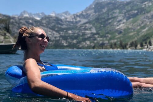 Person smiling and floating on an inflatable lounger in a lake, with mountains in the background on a sunny day. visit bishop