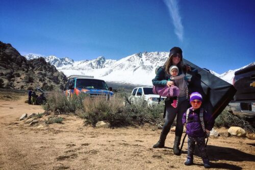 A woman with two kids stands in a mountainous area near parked vehicles under a clear blue sky. visit bishop