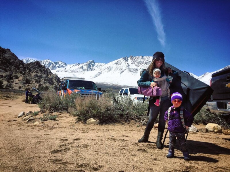 A woman with two kids stands in a mountainous area near parked vehicles under a clear blue sky. visit bishop