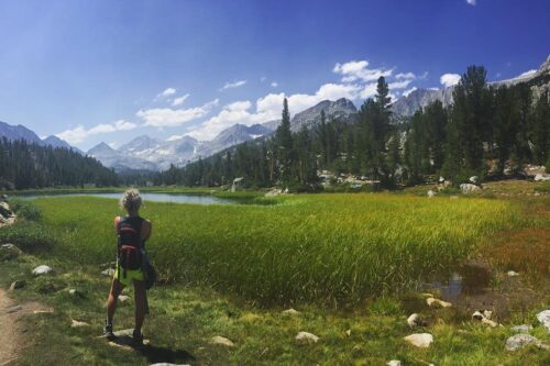 Person with a backpack stands on a trail, looking at a serene mountain landscape with a lake and lush greenery. visit bishop