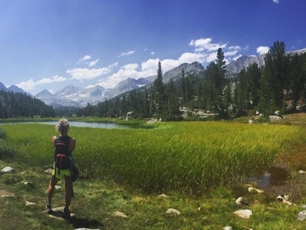 Person with a backpack stands on a trail, looking at a serene mountain landscape with a lake and lush greenery. visit bishop