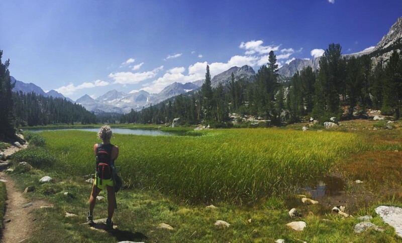 Person with a backpack stands on a trail, looking at a serene mountain landscape with a lake and lush greenery. visit bishop