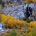 House beside a rocky cliff surrounded by vibrant autumn trees with a small waterfall cascading down the rocks, reminiscent of the serene landscapes near Bishop, California. visit bishop