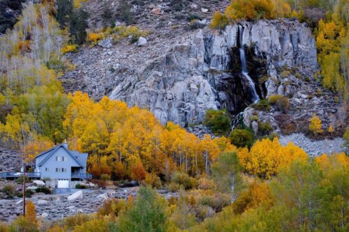 House beside a rocky cliff surrounded by vibrant autumn trees with a small waterfall cascading down the rocks, reminiscent of the serene landscapes near Bishop, California. visit bishop