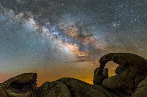 A stunning night sky featuring the Milky Way galaxy with vibrant stars and celestial colors. In the foreground, unique, weathered rock formations in Bishop, California, are silhouetted against the brilliant backdrop of stars. visit bishop