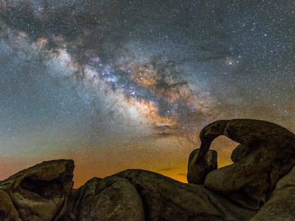 A stunning night sky featuring the Milky Way galaxy with vibrant stars and celestial colors. In the foreground, unique, weathered rock formations in Bishop, California, are silhouetted against the brilliant backdrop of stars. visit bishop