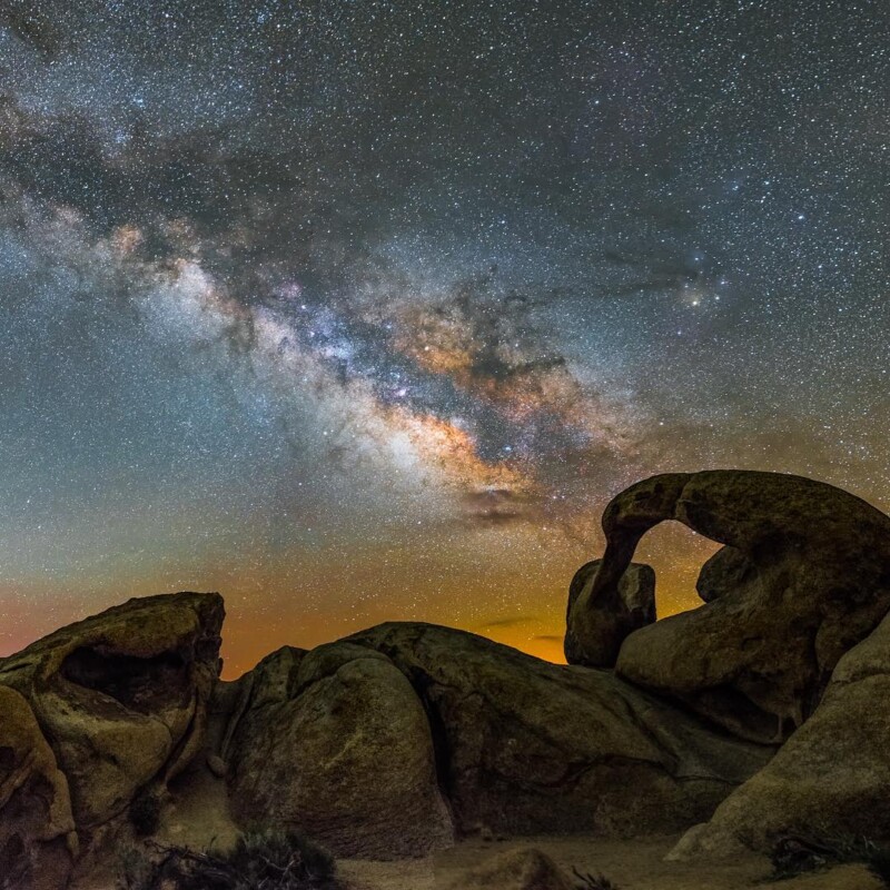 A stunning night sky featuring the Milky Way galaxy with vibrant stars and celestial colors. In the foreground, unique, weathered rock formations in Bishop, California, are silhouetted against the brilliant backdrop of stars. visit bishop