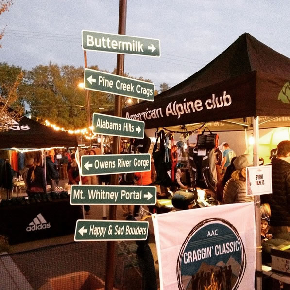 Outdoor market scene with signs pointing to various locations, and a booth displaying "American Alpine Club" and "Craggin' Classic" banners. visit bishop