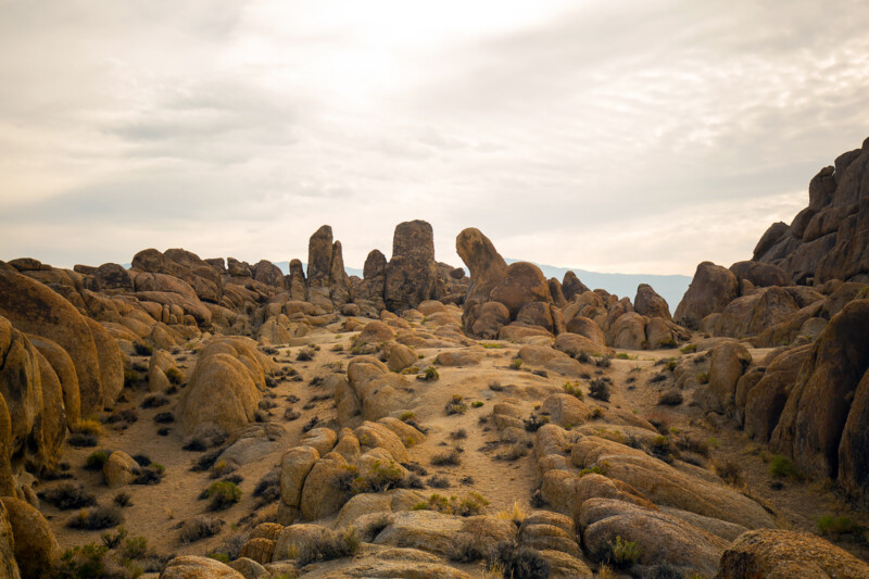 A rugged desert landscape with large rock formations under a cloudy sky. Sparse vegetation dots the sandy ground. visit bishop