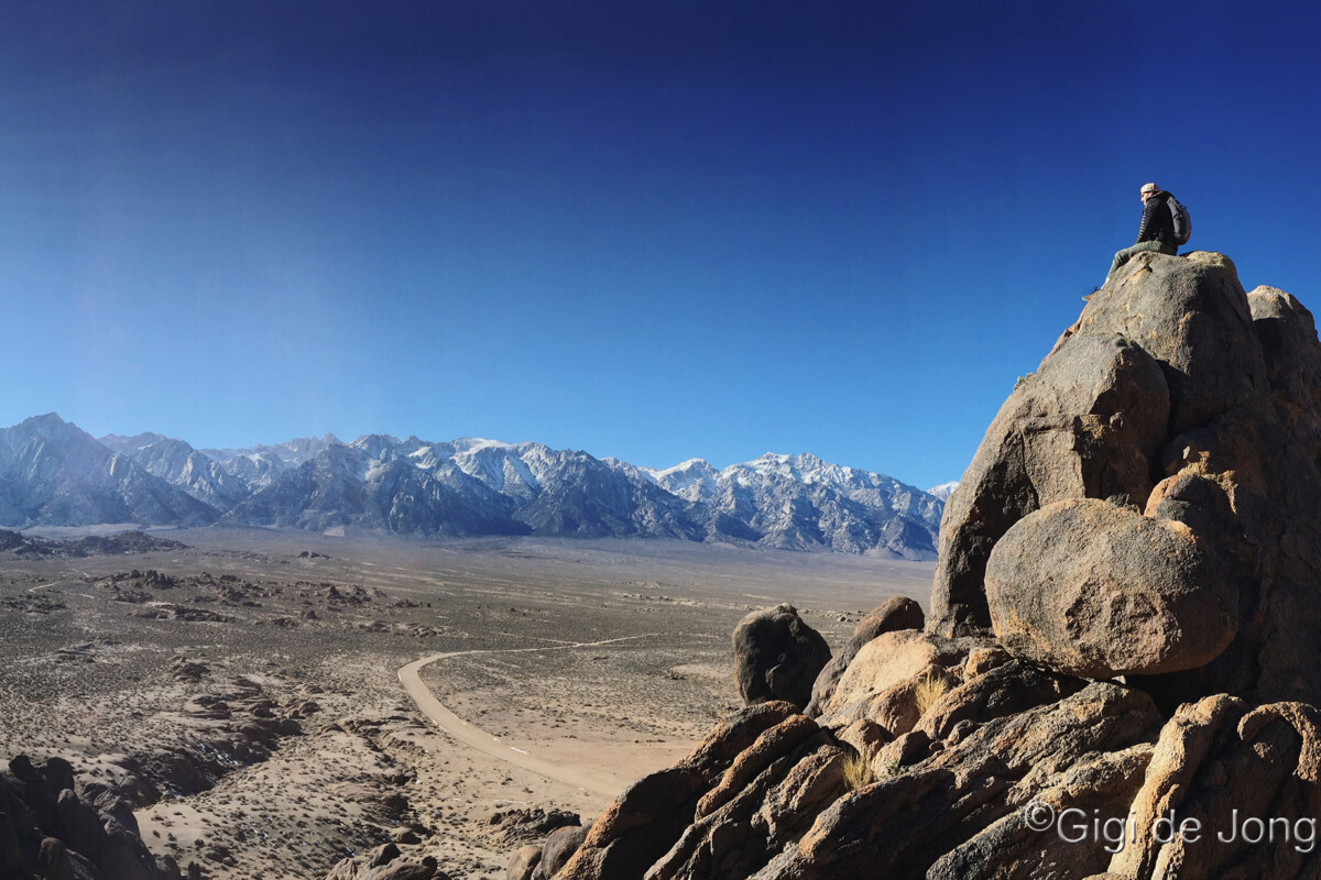 Person sitting on large rocks overlooking a vast desert landscape with distant mountains under a clear blue sky. visit bishop