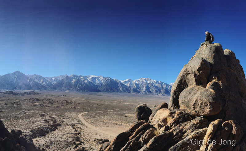 Person sitting on large rocks overlooking a vast desert landscape with distant mountains under a clear blue sky. visit bishop