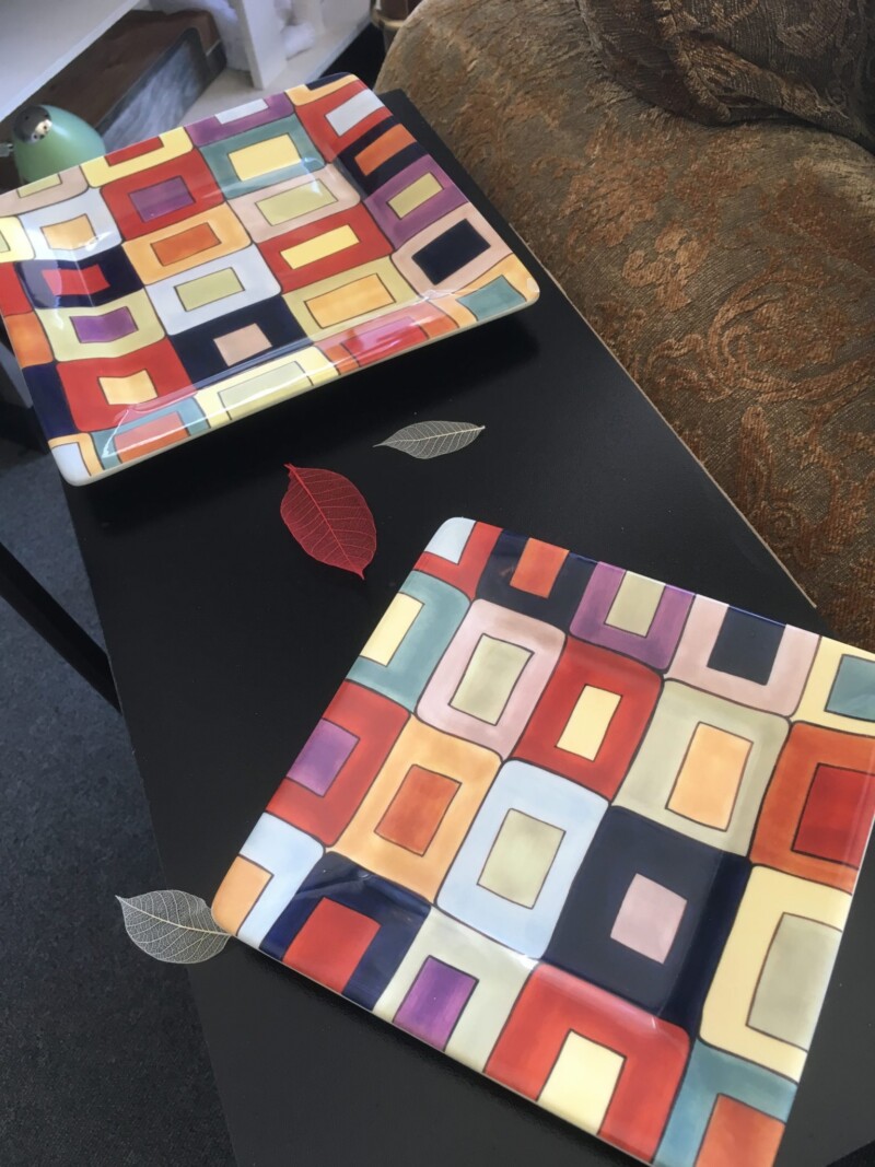 Two colorful, square plates with geometric designs on a black table, next to a patterned brown sofa. visit bishop