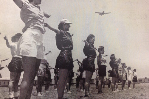 A group of women stand in a line doing exercises outdoors while an airplane flies overhead. visit bishop
