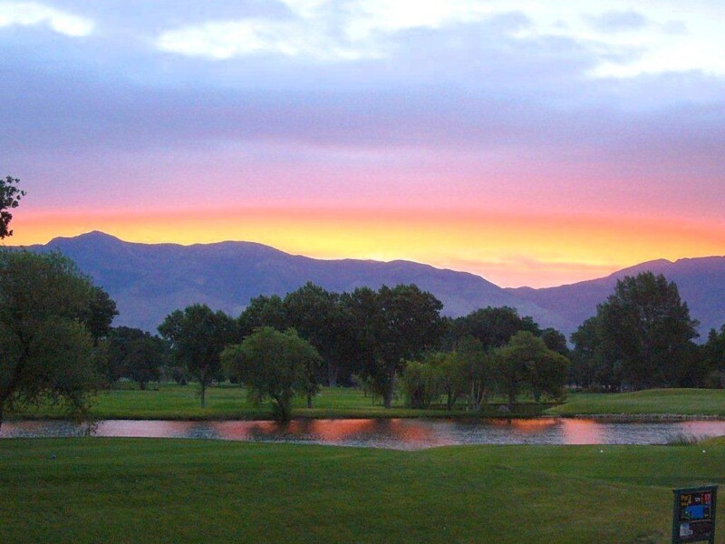 A serene landscape featuring a vibrant sunset over the distant mountains of Bishop, California. The sky showcases hues of orange, pink, and purple. Below, a peaceful lake reflects the colorful sky, surrounded by lush green trees and grass. visit bishop