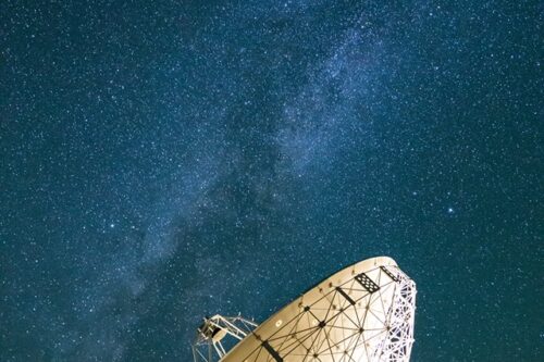 A large satellite dish in Bishop, California is pointed towards the starry night sky, with the Milky Way galaxy clearly visible above it. The dish is part of a ground station installation, with parts of other structures visible at the bottom of the image. visit bishop