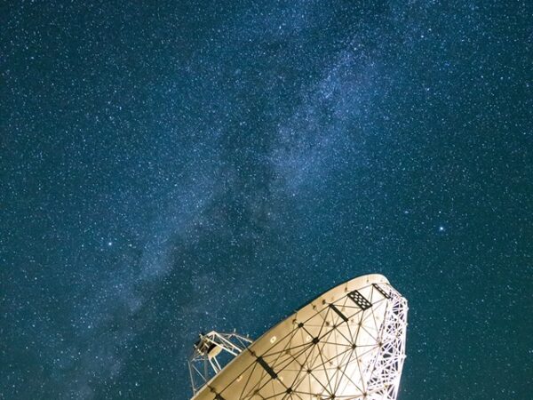A large satellite dish in Bishop, California is pointed towards the starry night sky, with the Milky Way galaxy clearly visible above it. The dish is part of a ground station installation, with parts of other structures visible at the bottom of the image. visit bishop