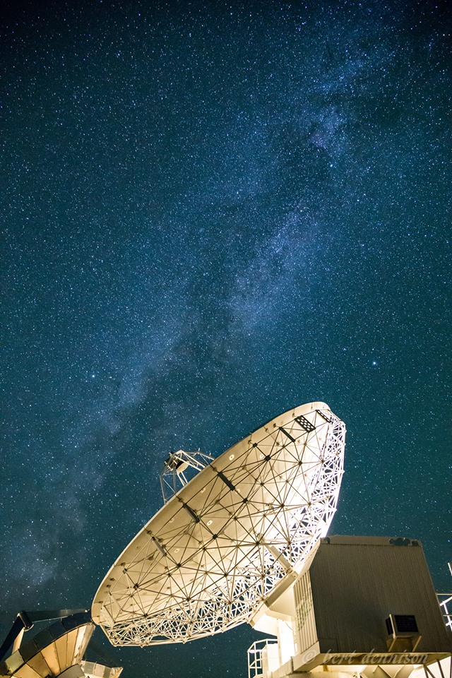 A large satellite dish in Bishop, California is pointed towards the starry night sky, with the Milky Way galaxy clearly visible above it. The dish is part of a ground station installation, with parts of other structures visible at the bottom of the image. visit bishop