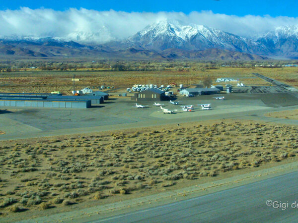 Aerial view of a small airfield with several planes, hangars, and buildings, with snow-capped mountains in the background. visit bishop