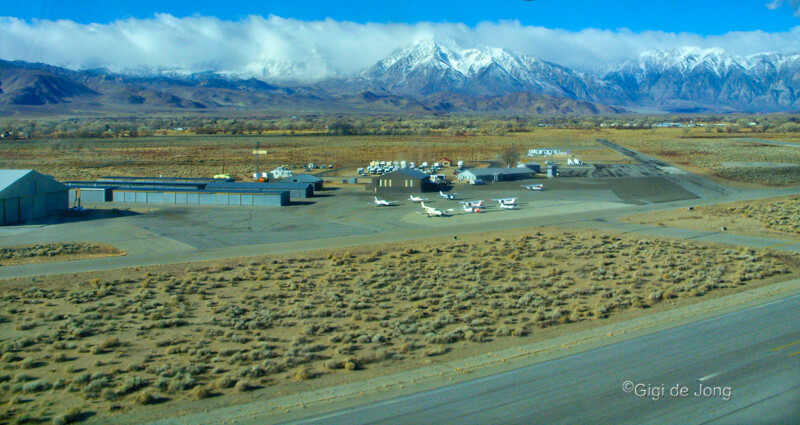 Aerial view of a small airfield with several planes, hangars, and buildings, with snow-capped mountains in the background. visit bishop