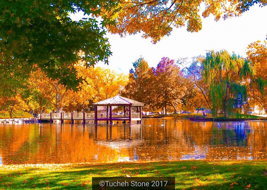 A serene park in Bishop, California, with vibrant autumn foliage surrounds a calm lake. In the center, a wooden gazebo sits on the water, connected by a bridge. The leaves on the trees display brilliant shades of orange, yellow, and green, reflecting beautifully on the lake's surface. visit bishop
