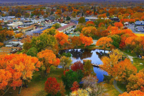 Aerial view of Bishop, California, surrounded by autumn foliage with vibrant orange, red, and yellow trees. A reflective pond is situated in the middle of a park. Snow-capped mountains and a partly cloudy sky are visible in the background. visit bishop