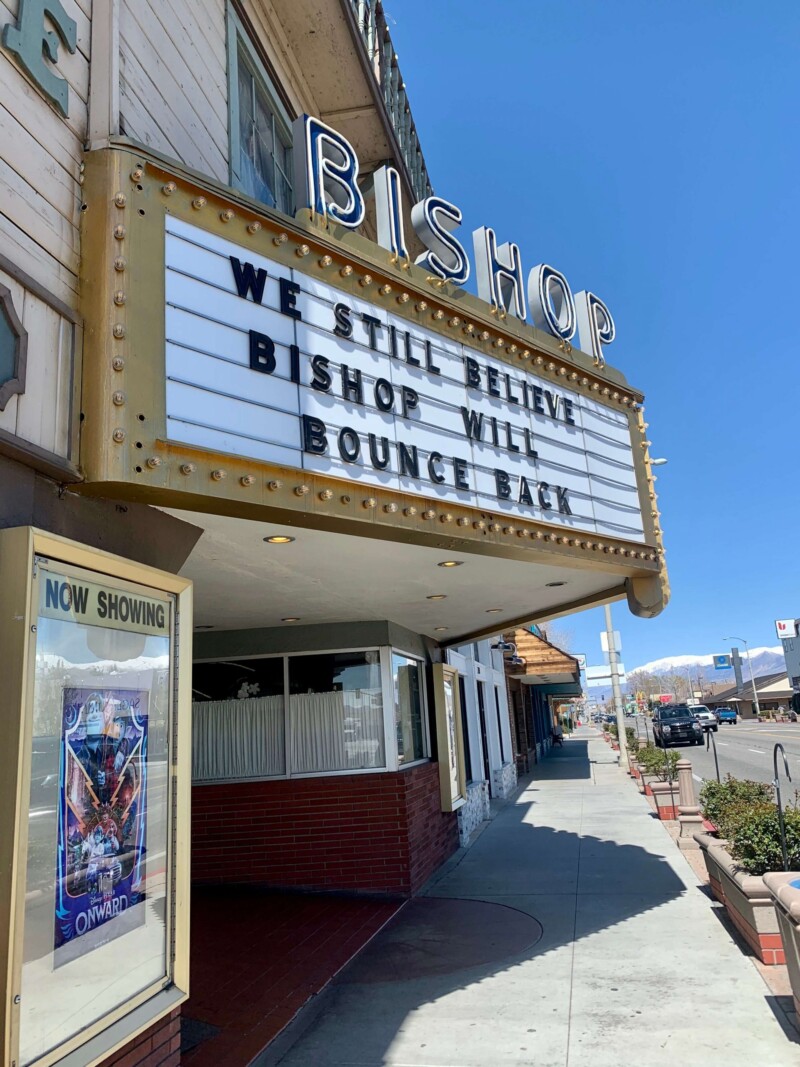 A vintage cinema marquee reads "We still believe Bishop will bounce back." The theater's sign above the marquee says "Bishop," and a poster for the movie "Onward" is visible in a display case to the left of the entrance. On this bright, sunny day in Bishop, California, optimism fills the air. visit bishop