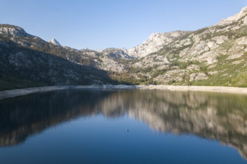 A serene lake in Bishop, California reflects the surrounding mountains under a clear blue sky. The rugged peaks are partially covered with vegetation. A lone kayaker paddles on the calm water in the center of the lake, adding a sense of scale and tranquility to the scene. visit bishop