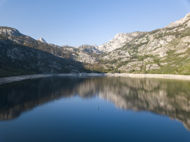 A serene lake in Bishop, California reflects the surrounding mountains under a clear blue sky. The rugged peaks are partially covered with vegetation. A lone kayaker paddles on the calm water in the center of the lake, adding a sense of scale and tranquility to the scene. visit bishop