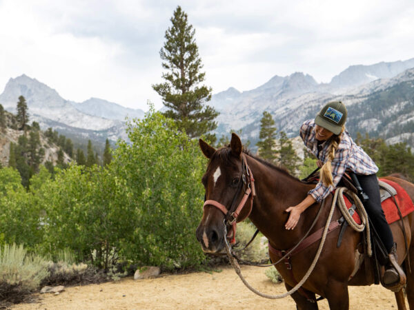A person wearing a plaid shirt and baseball cap sits on a brown horse, patting its neck. They are outdoors surrounded by lush green trees and shrubs, with a mountain range in the background under a mildly cloudy sky in Bishop, California. visit bishop